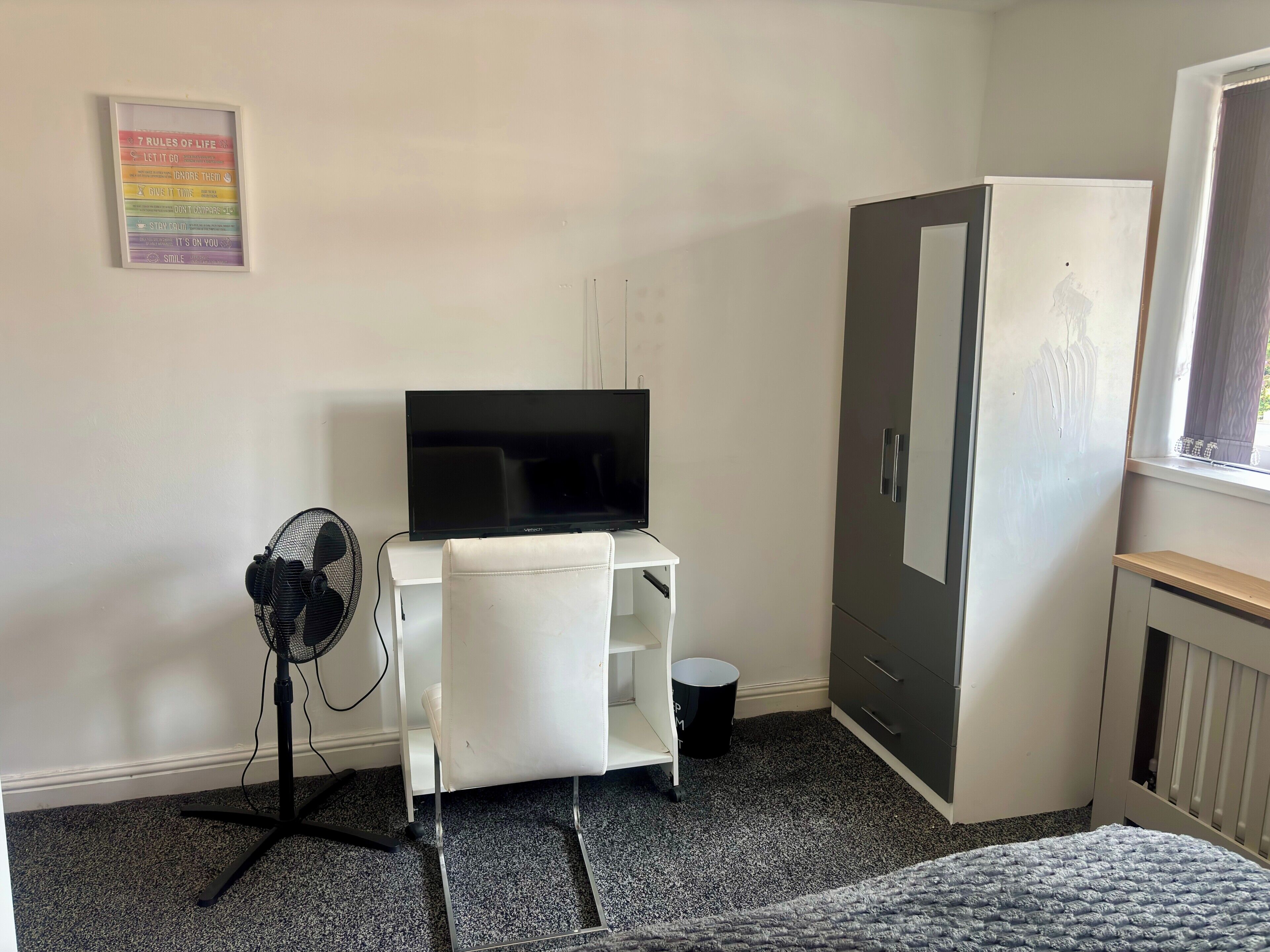 Ein Zimmer mit Schreibtisch, Fernseher, Ventilator, Kleiderschrank und Fenster.