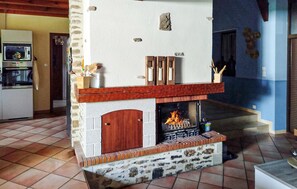 Ein Kamin aus Stein mit hölzernem Sims, ein Fliesenboden und ein Küchenbereich mit Geräten.