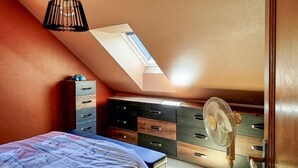 Ein Schlafzimmer mit schrägem Dach, einem Bett, einer Kommode und einem Standventilator.