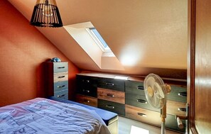 Ein Schlafzimmer mit schrägem Dach, einem Bett, einer Kommode und einem Standventilator.
