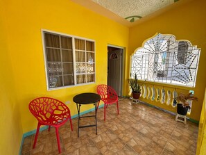 A room with yellow walls, a tiled floor, a window with a metal grill, a door, and two red chairs with a small table. - Mammee Bay Hideaway (St. Ann Parish)