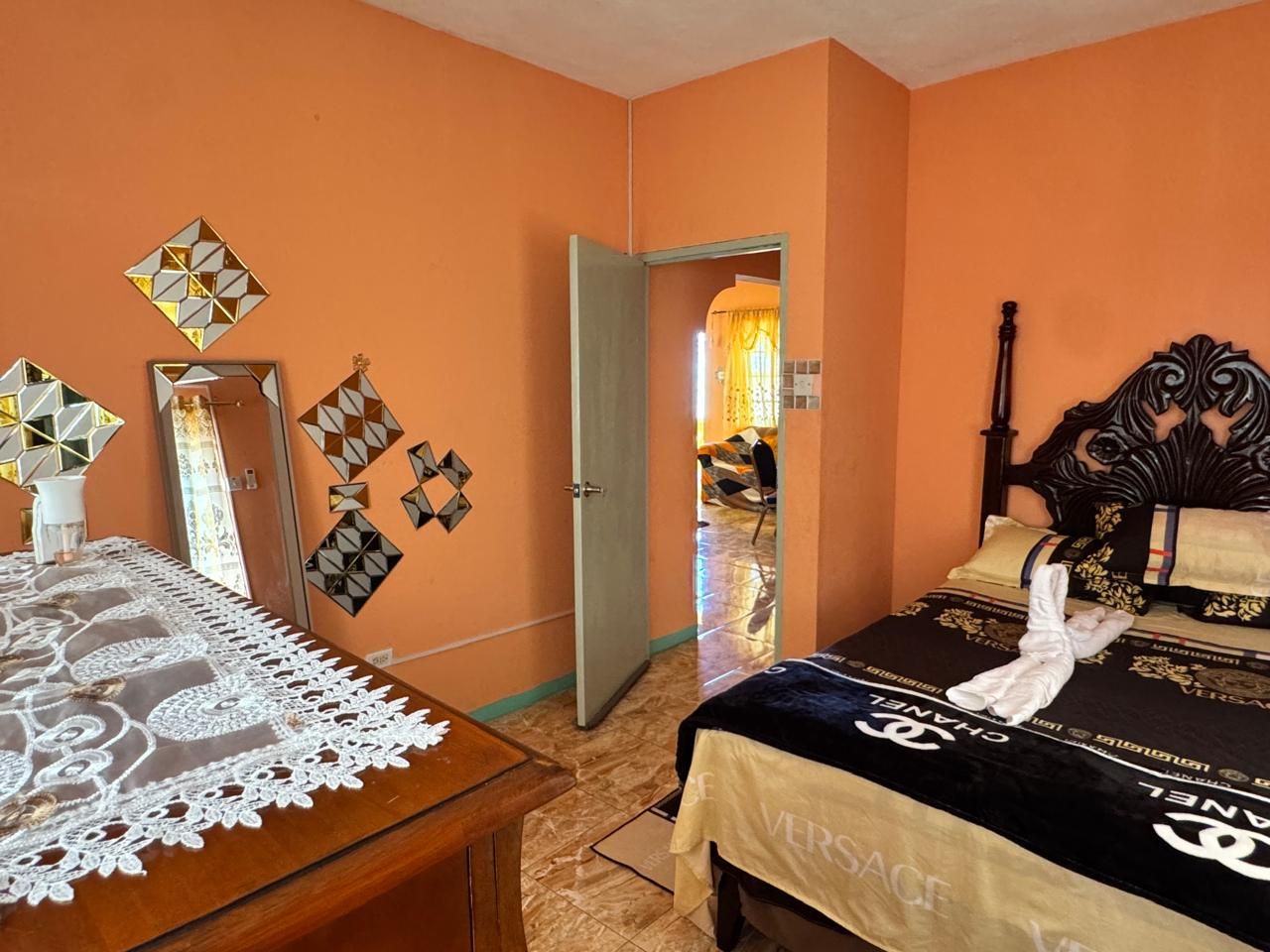 A hotel room with a bed, a wooden nightstand, a mirror, and a door leading to another room.