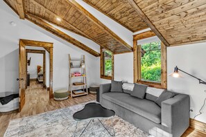 A modern living room with a grey sofa, wooden beams, a ladder, and a view of greenery. - Vintage Vale at Eagles Nest - Views, yard games, hot tub, outdoor fireplace (Elk Park)