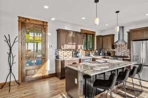A modern kitchen with wooden cabinets, a large island with bar seating, and a view through a glass door. - Vintage Vale at Eagles Nest - Views, yard games, hot tub, outdoor fireplace (Elk Park)