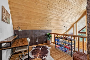 Ein gemütliches Zimmer mit schrägem Dach, einem Schreibtisch mit Lampe, einem Kuhfellteppich und einem Regal mit Gesellschaftsspielen.