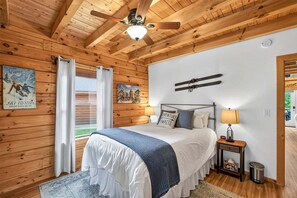 A wooden-paneled bedroom with a bed, a ceiling fan, a window with curtains, a wall-mounted picture, and a bedside table with a lamp. - White Fir Cabin | Hot Tub | Fire Pit | Near Tamarack (Donnelly)