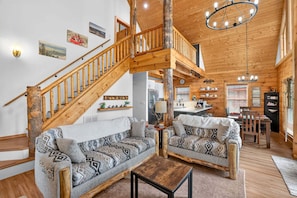 A cabin interior with a wooden staircase, two patterned sofas, a dining area, and a kitchen. - White Fir Cabin | Hot Tub | Fire Pit | Near Tamarack (Donnelly)