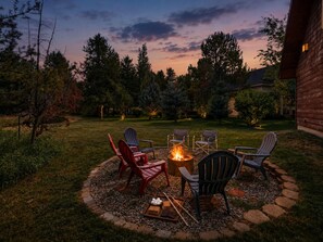 Un espace extérieur circulaire aménagé avec des sièges, comprenant un foyer, des chaises et une table, le tout situé dans un jardin avec des arbres et un bâtiment en arrière-plan.