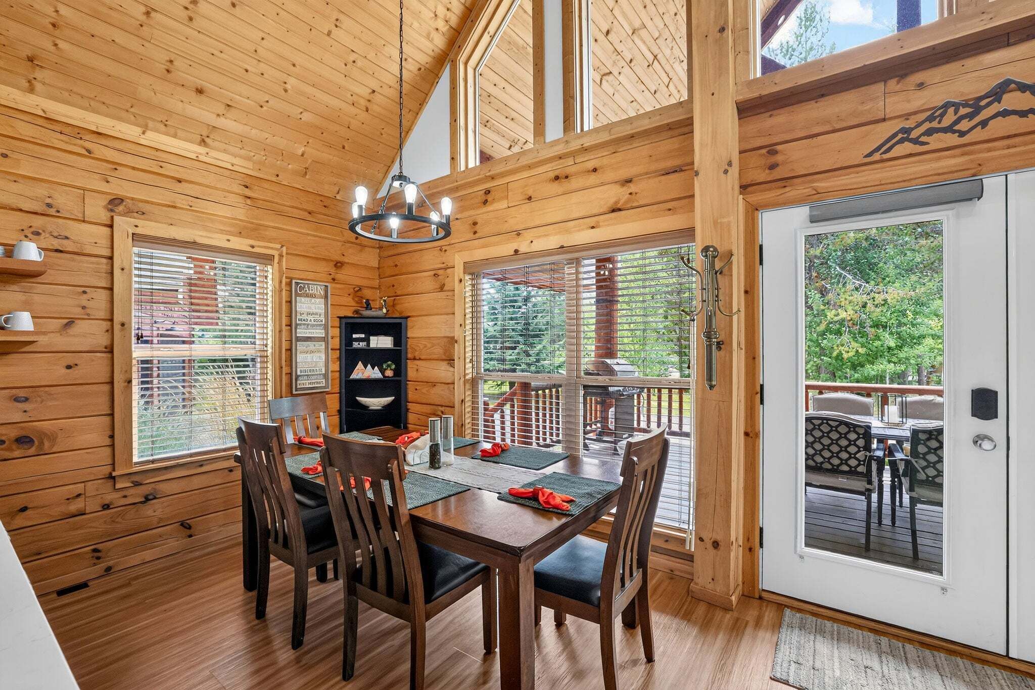 Interior de una cabaña de madera con una mesa de comedor lista para cuatro comensales, una araña y un letrero de cabaña en la pared.