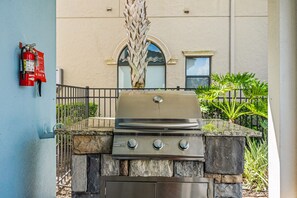 An outdoor grill with a stone base, a fire extinguisher on the wall, and a palm tree in the background. - Legends Coastal Cottage, Arcade, Golf simulator, Near Disney (Kissimmee)