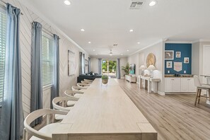 Une longue table à manger avec des chaises, un canapé et un coin cuisine équipé de placards et d’un plan de travail.