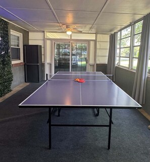 Ein Tischtennis-Tisch in einem Raum mit Deckenventilator, großen Fenstern und einer zur Außenseite führenden Tür.