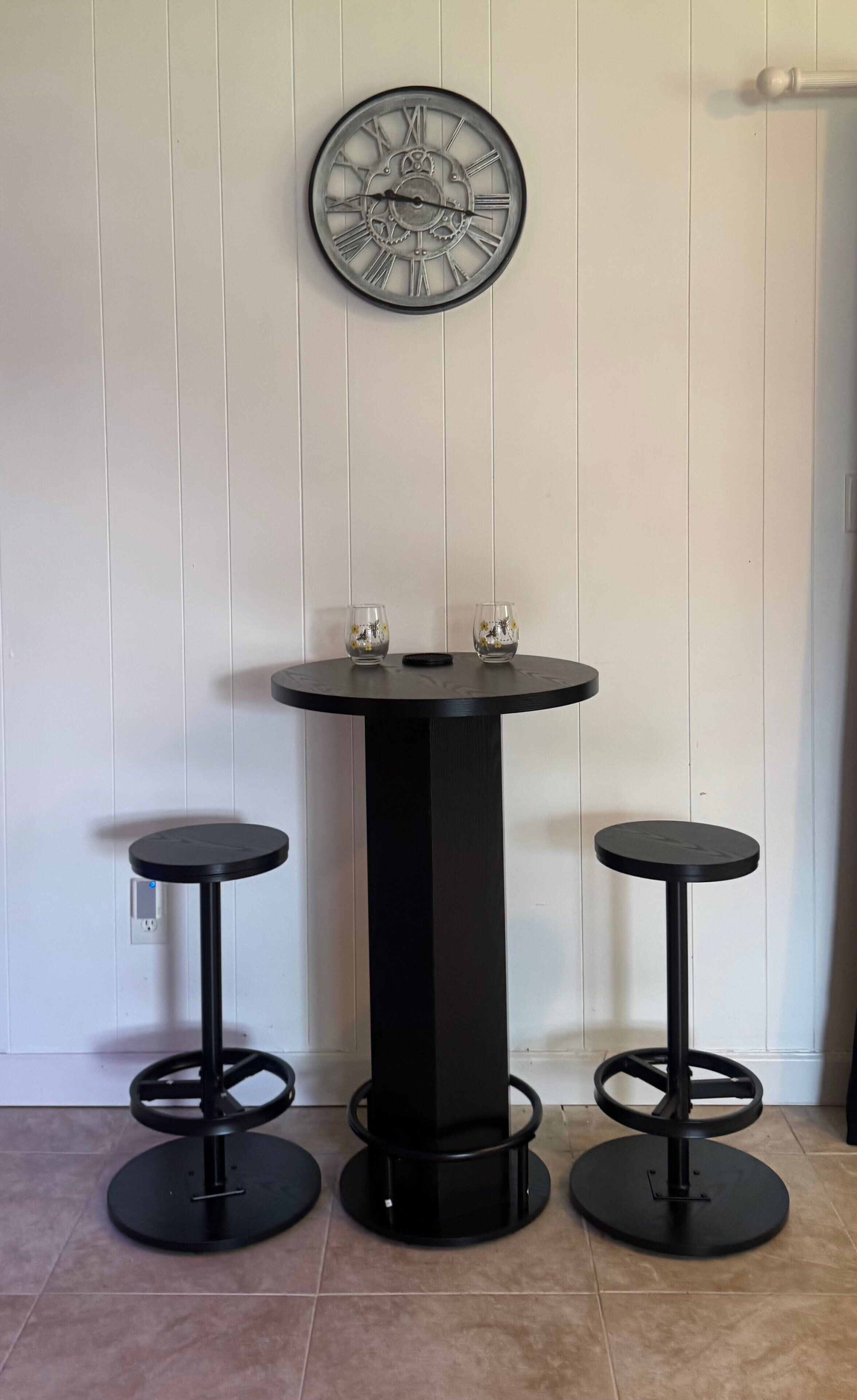 A black round table with two stools, a wall clock, and a power outlet.