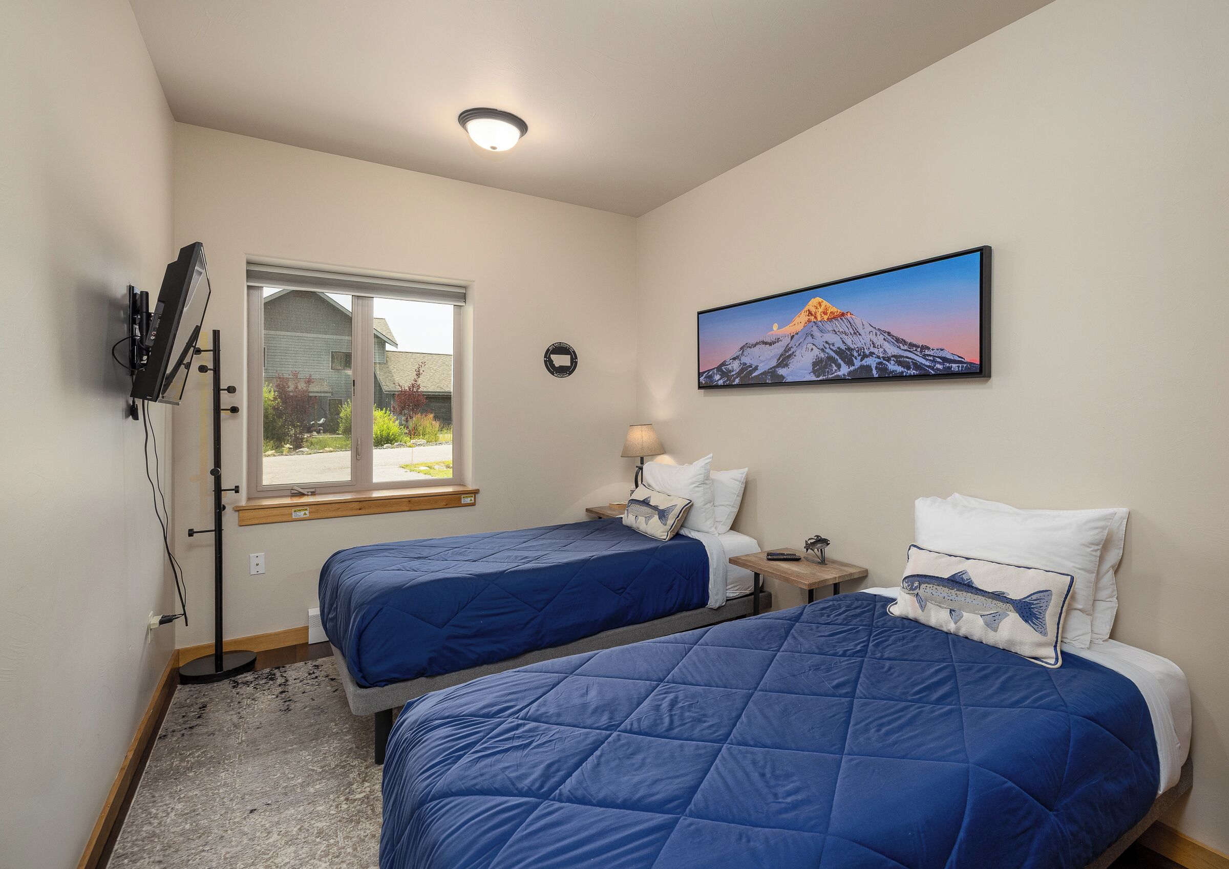A hotel room with two beds, a window with a view of a house, a painting of a mountain, and a ceiling light.