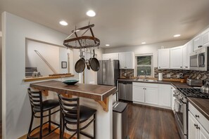 A modern kitchen with white cabinets, stainless steel appliances, a wooden island with bar stools, and a hanging pot rack. - New Listing! | Private Hot Tub & Minutes to Big Sky Resort and Town Center (Big Sky)