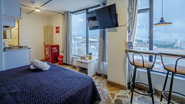 A hotel room with a bed, a desk with a chair, a television, and a window with a view of the city. - Coliving Majestic (Manizales)