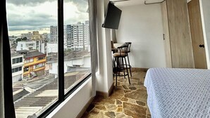 A room with a large window, a bed, a desk with a chair, and a television. - Coliving Majestic (Manizales)