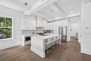 A modern kitchen with white cabinetry, a central island with bar stools, stainless steel appliances, and a view of greenery outside. - Easy Walk 2 Beach! Private Pool, LSV/Golf Cart & Bikes! (Santa Rosa Beach)