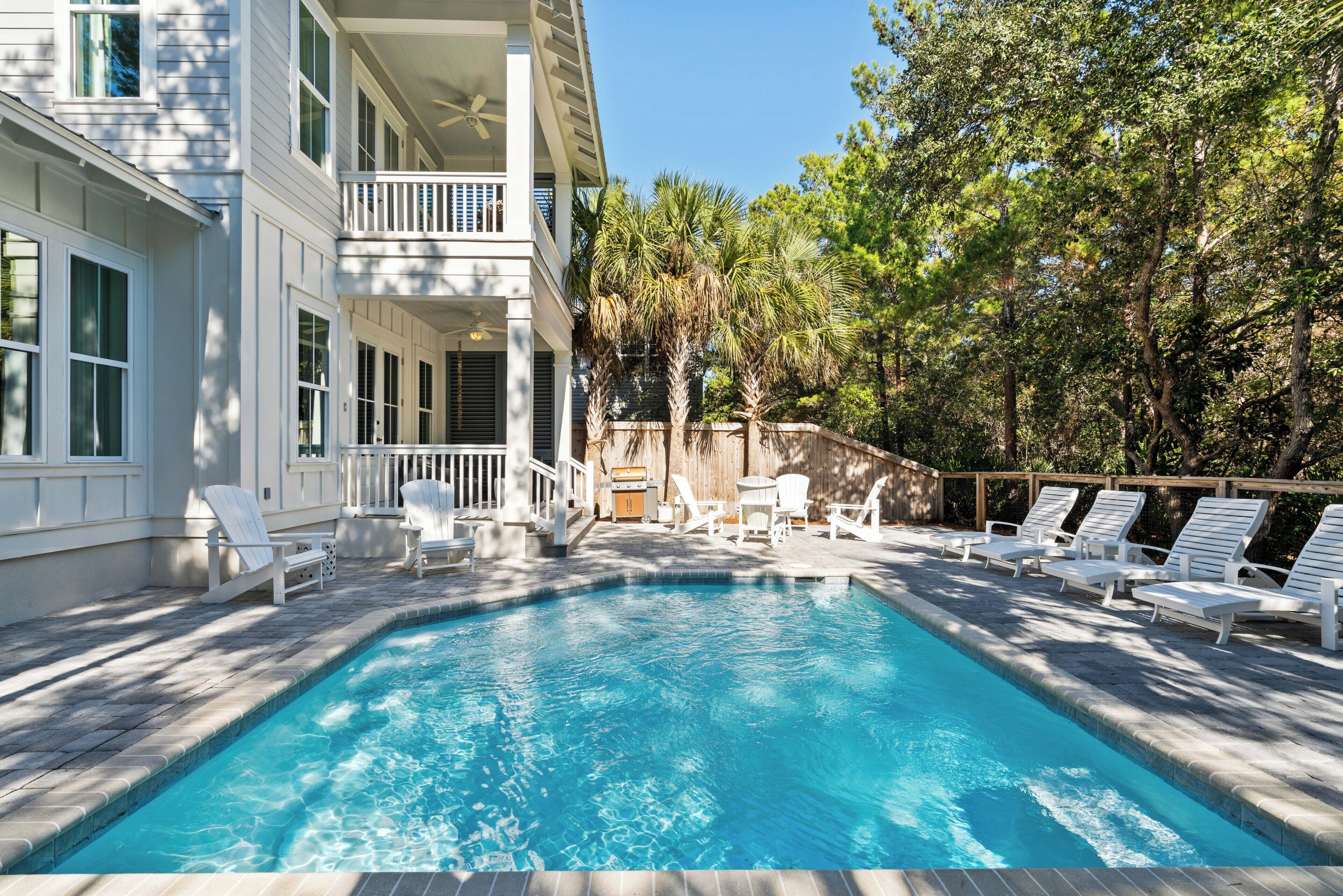 A swimming pool with lounge chairs and a barbecue area, surrounded by trees and a building with balconies.