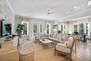 A spacious living room with a sectional sofa, a coffee table, and a dining area with a chandelier. - NEW! EZ Walk to Beach! Private Pool, LSV/Golf Cart, & Bikes! (Santa Rosa Beach)