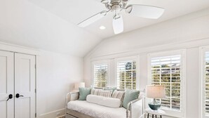 Ein Schlafzimmer mit einem Bett, einem Deckenventilator, einem Fenster mit Jalousien, einem kleinen Tisch mit einer Lampe und einem gemusterten Teppich.