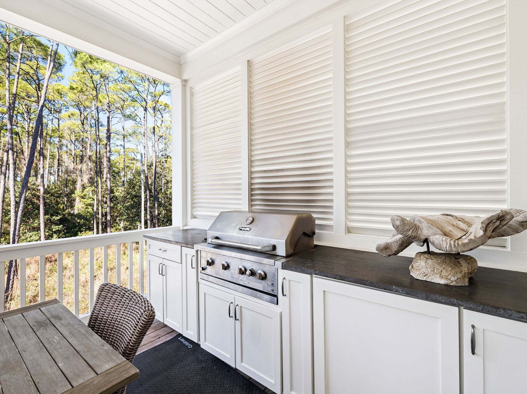 A covered outdoor kitchen with a grill, white cabinetry, and a view of trees.