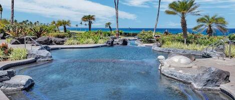 A swimming pool with a curved design, surrounded by rocks and palm trees, with a view of the ocean.