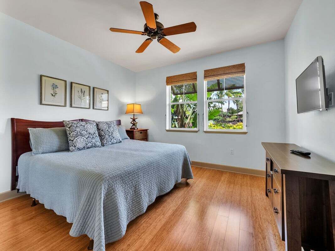 A bedroom with a bed, a ceiling fan, a nightstand, a lamp, a TV, and windows with blinds.