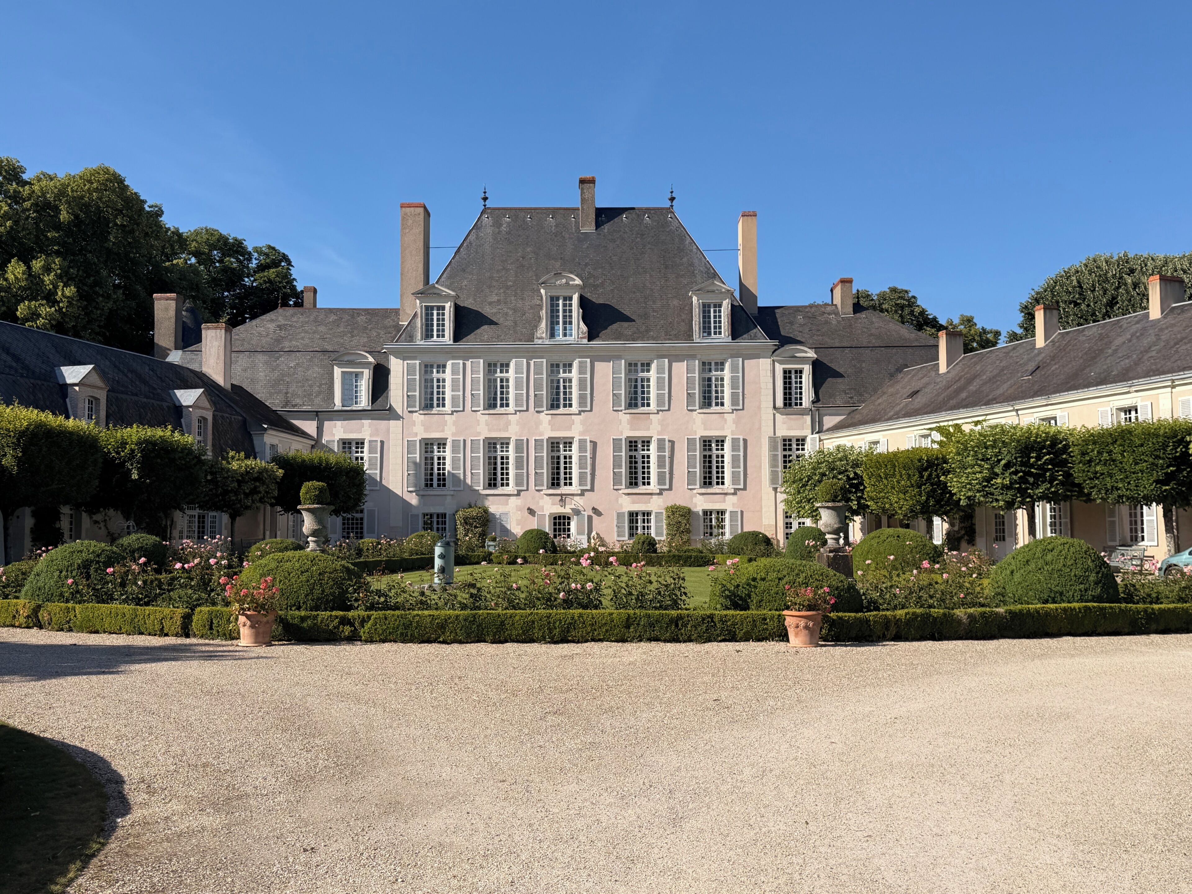 A large, historic building with multiple chimneys, a symmetrical facade, and a well-maintained garden with hedges and flower beds.