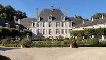 Un grand bâtiment historique, doté de nombreuses cheminées, d’une façade symétrique et d’un jardin bien entretenu, agrémenté de haies et de parterres de fleurs.