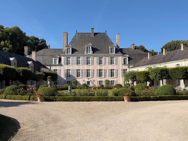 A large, historic building with multiple chimneys, a symmetrical facade, and a well-maintained garden with hedges and flower beds.