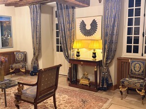 A room with a wooden chair, a table with two lamps, a decorative mirror, and curtains with a pattern.