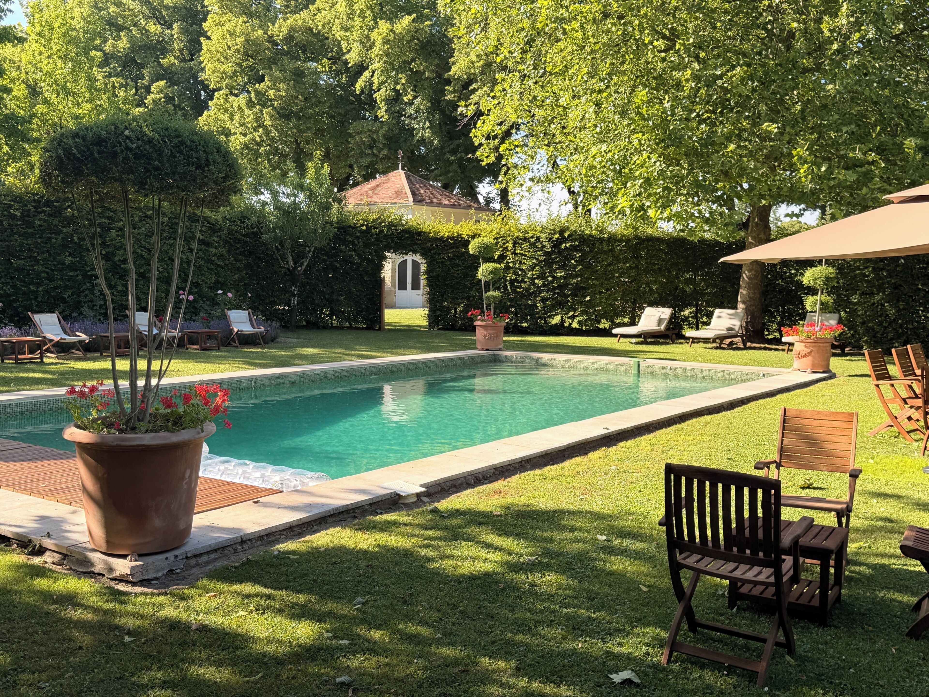 A swimming pool with a tiled deck, surrounded by grass and trees. There are lounge chairs and a potted plant.