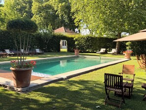 A swimming pool with a tiled deck, surrounded by grass and trees. There are lounge chairs and a potted plant.