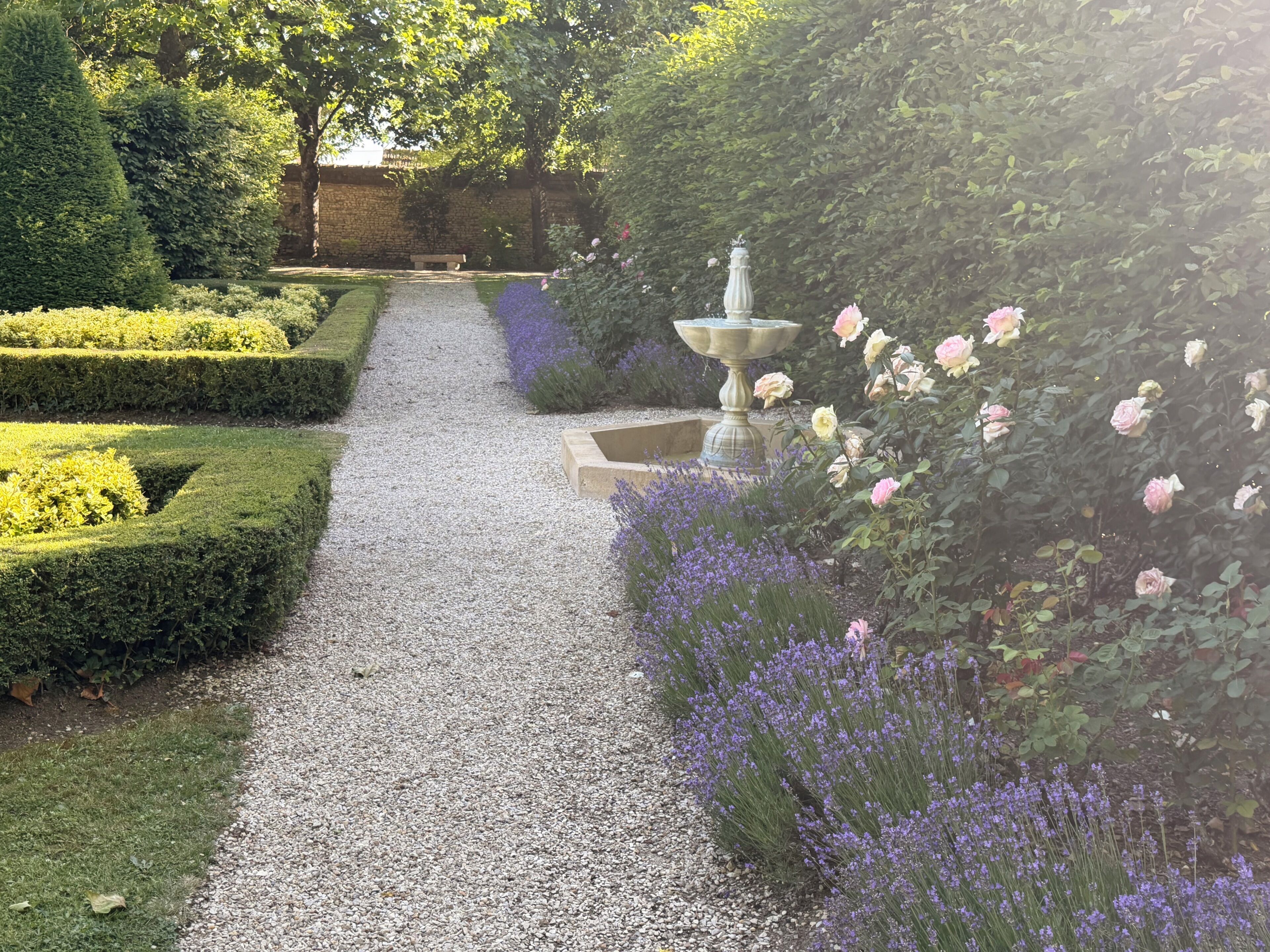 Un chemin de jardin avec une allée centrale en gravier, bordée de haies soigneusement taillées et de plantes en fleurs.