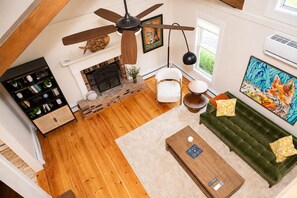 Een ruime woonkamer met een open haard, een plafondventilator, een groene bank met decoratieve kussens, en een boekenplank.