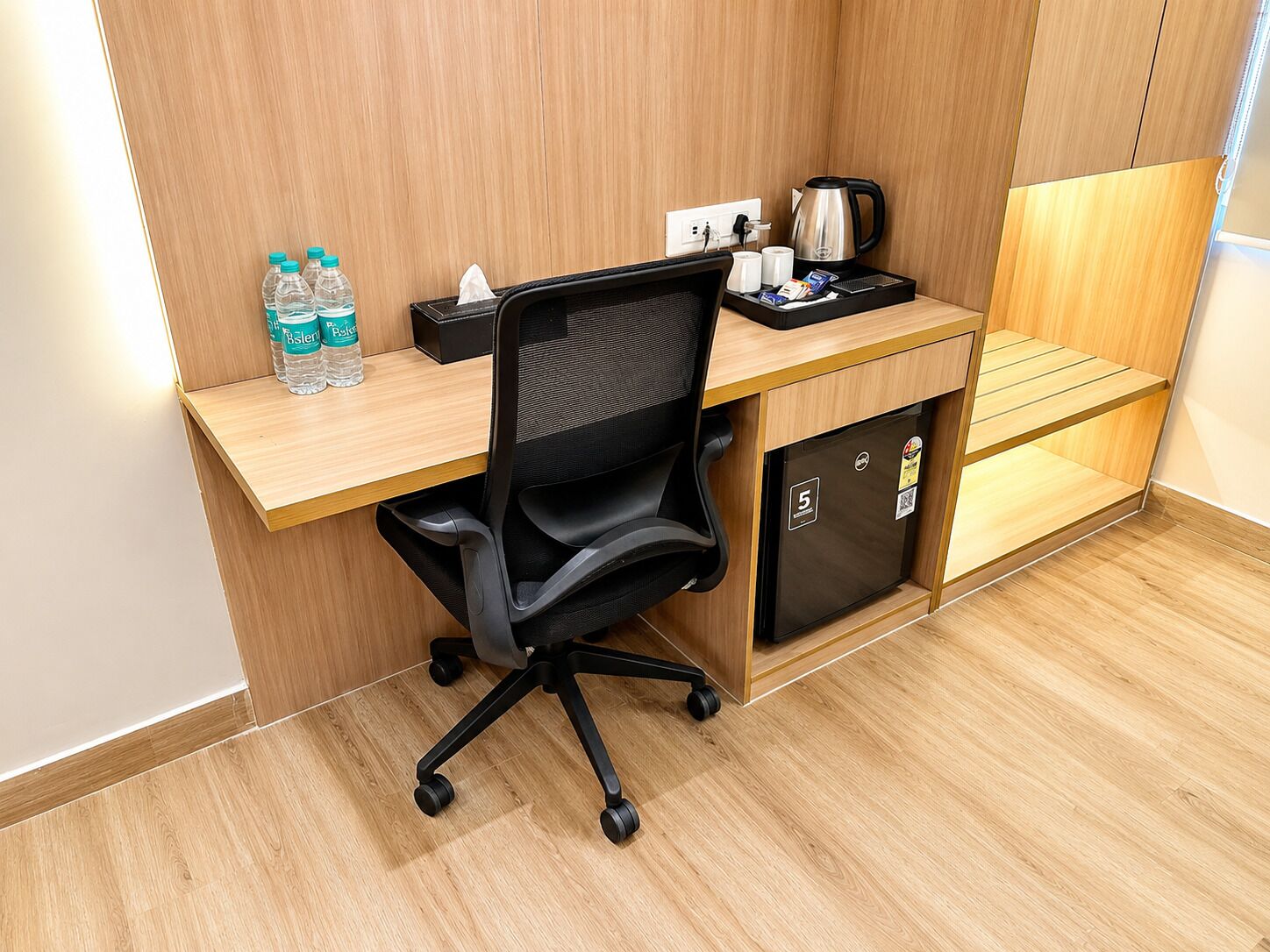 Espaço de escritório compacto, com mesa, cadeira, garrafas de água, cafeteira e mini geladeira.