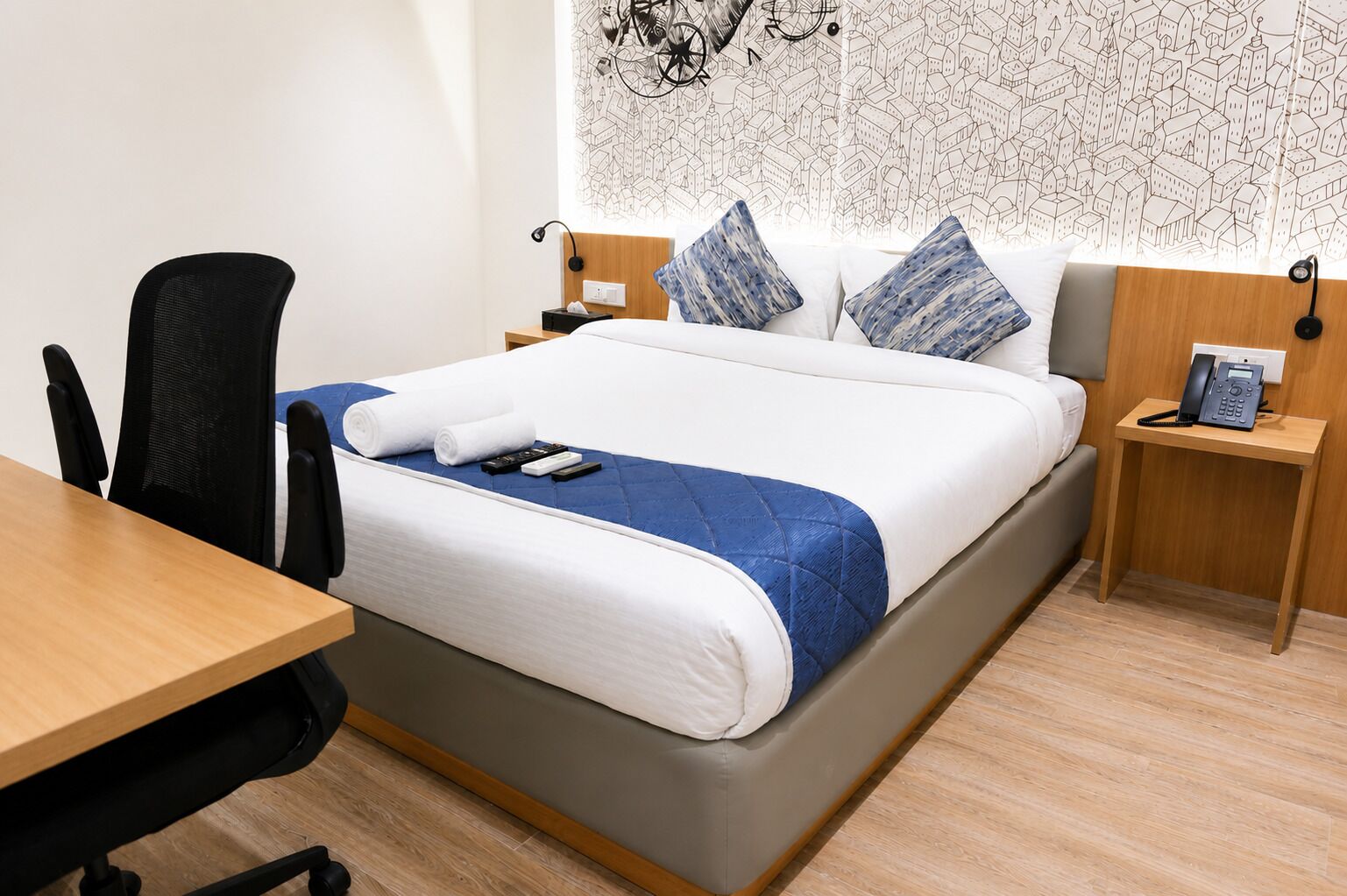Una camera d'hotel con un letto, una scrivania, una sedia, un telefono e una parete decorata con un motivo urbano.