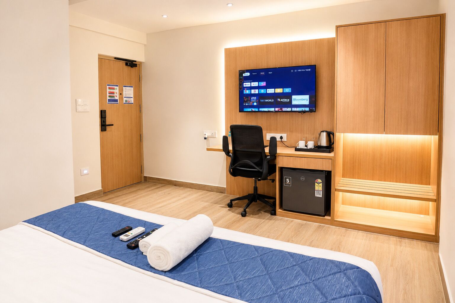 A hotel room with a bed, a desk with a computer, a chair, a small refrigerator, and a television on the wall.