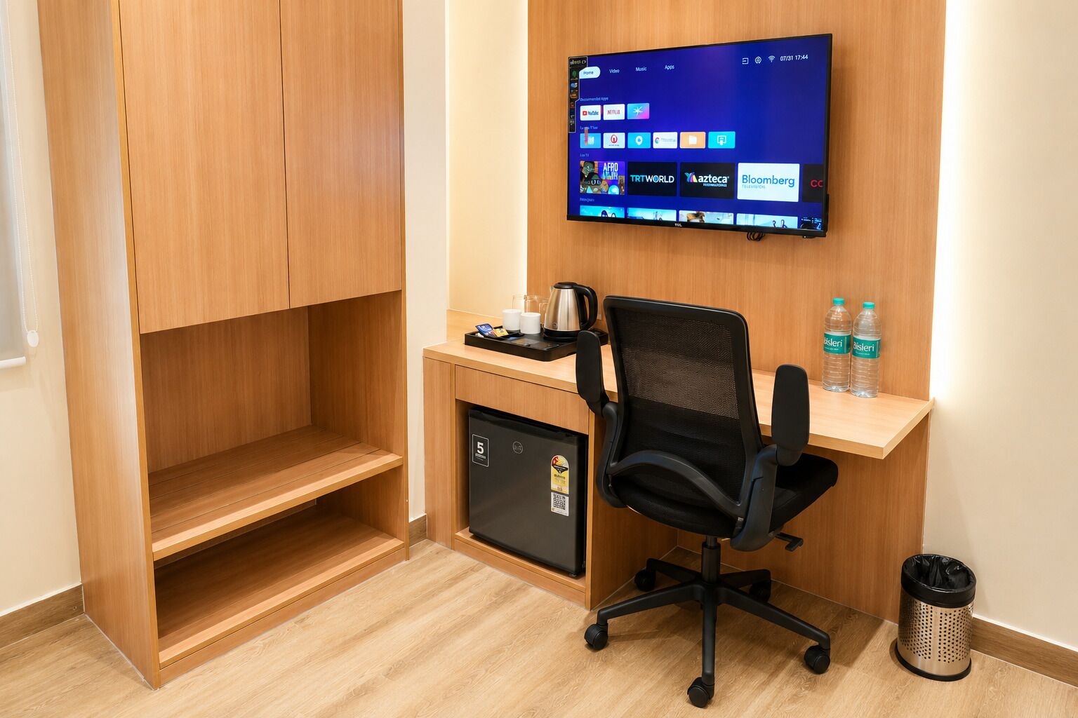 A hotel room with a wooden wardrobe, a desk with a computer monitor, a black office chair, a coffee maker, and a trash bin.