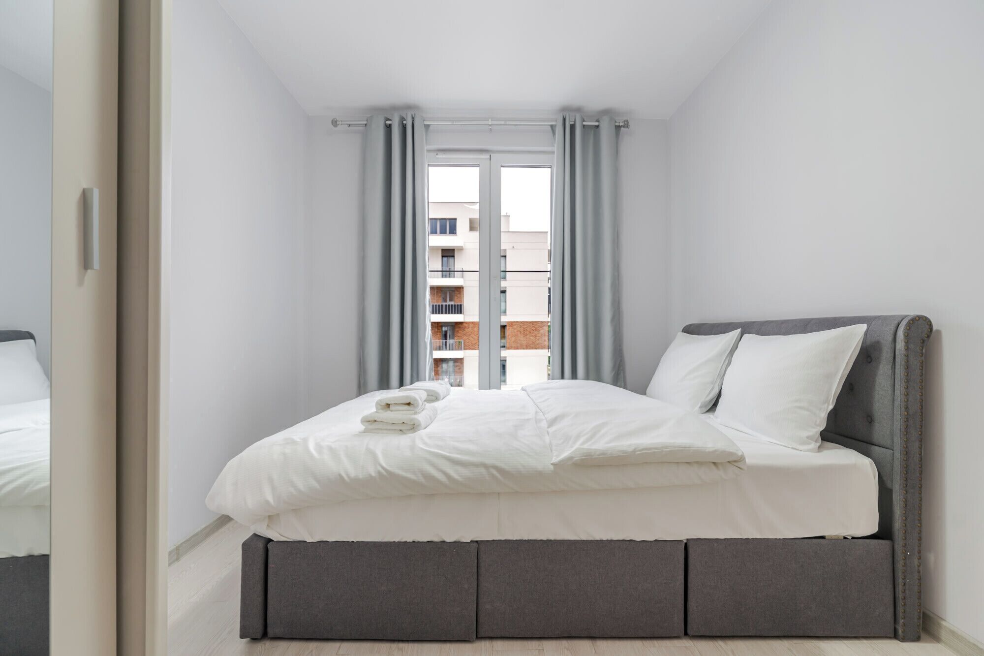 Un dormitorio moderno con una cama, cabecera gris y vista a un edificio a través de una ventana con cortinas grises.
