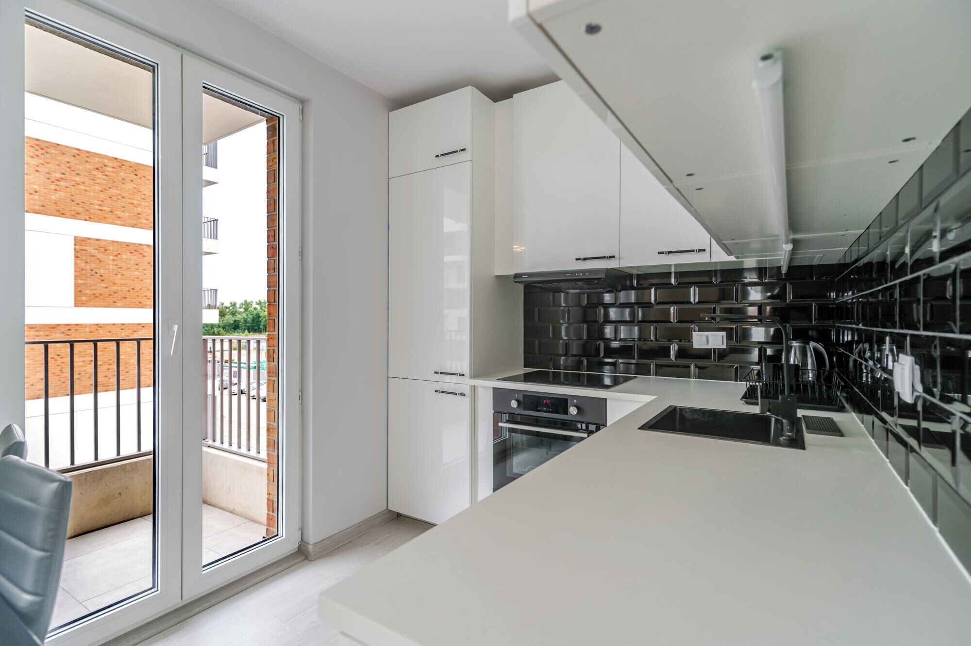 Una cocina moderna con armarios blancos, un protector contra salpicaduras negro y un ventanal corredero que da a un balcón.