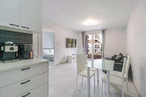 A modern kitchen with a coffee machine, a dining area with a glass table and chairs, and a view of a balcony with a building in the background. - Harmony Park Modern Apartment (Chorzów)