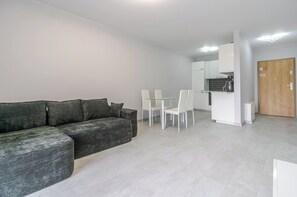 A modern living room with a grey sofa, a glass dining table with white chairs, and a kitchen area with white cabinets and a wooden door. - Harmony Park Modern Apartment (Chorzów)