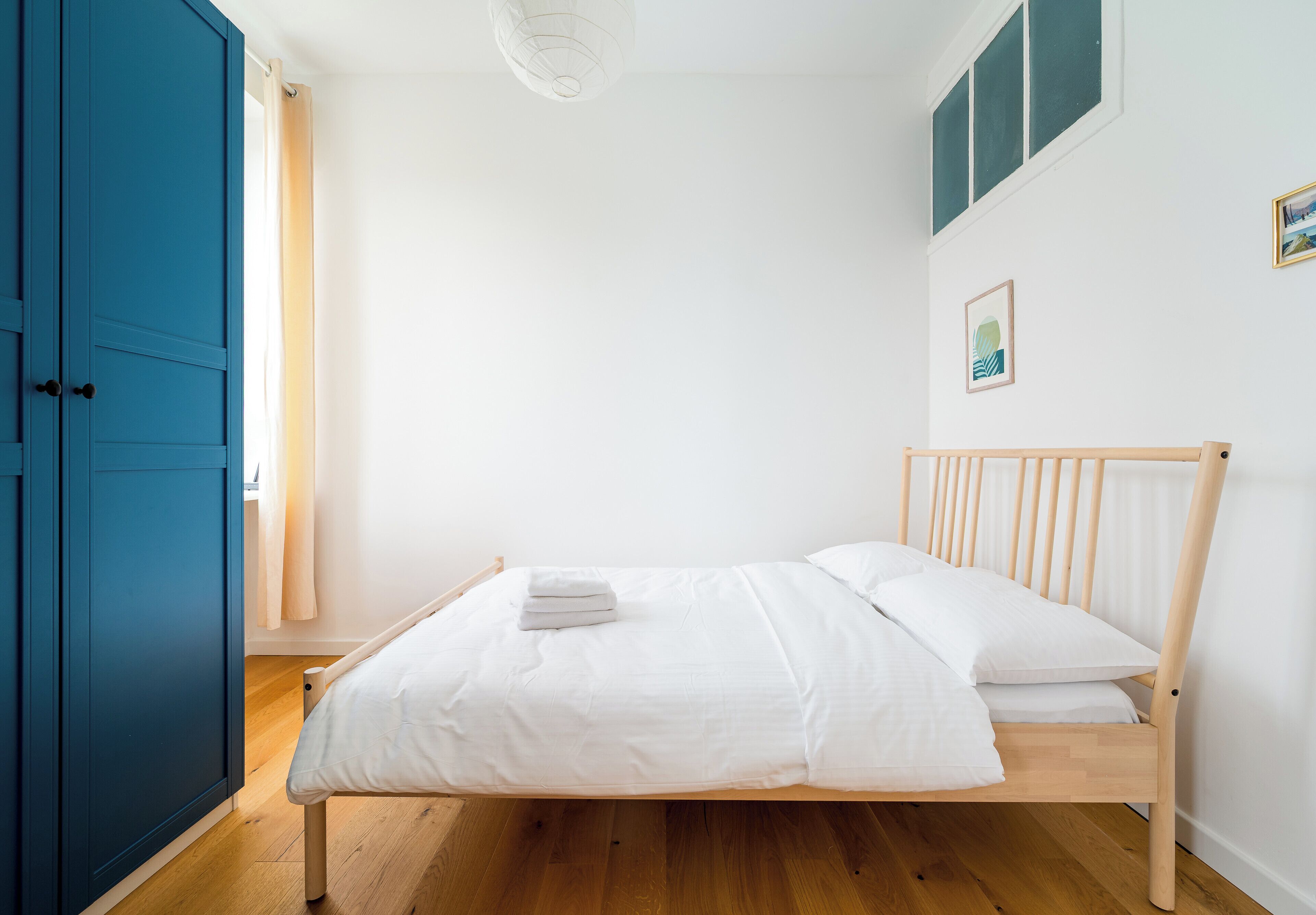 A single bed with a wooden frame and white bedding in a room with a blue wardrobe and a window with a curtain.