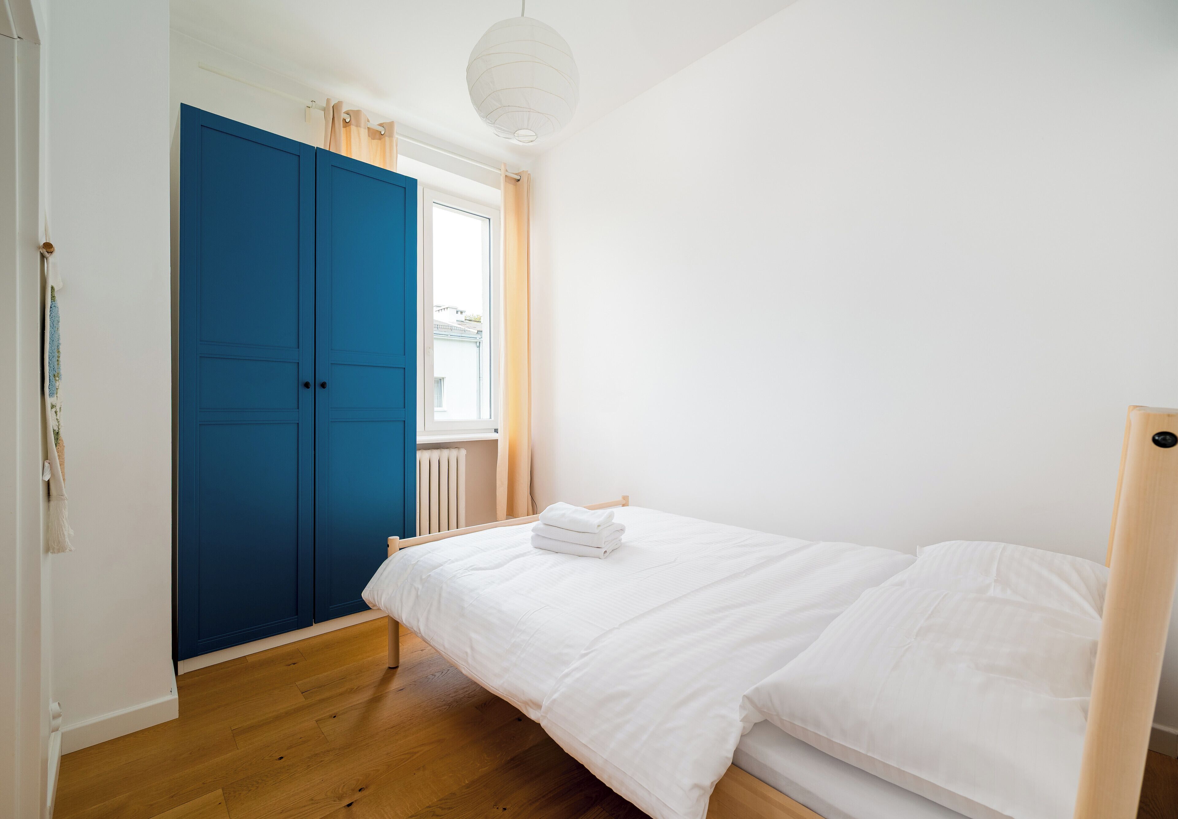 Une chambre avec un lit simple, une armoire bleue et une fenêtre avec des rideaux.