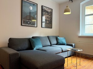 Ein modernes Wohnzimmer mit einem grauen Sofa, Türkisen Kissen, einem kleinen runden Tisch mit einer Fernbedienung und zwei gerahmten Bildern an der Wand.