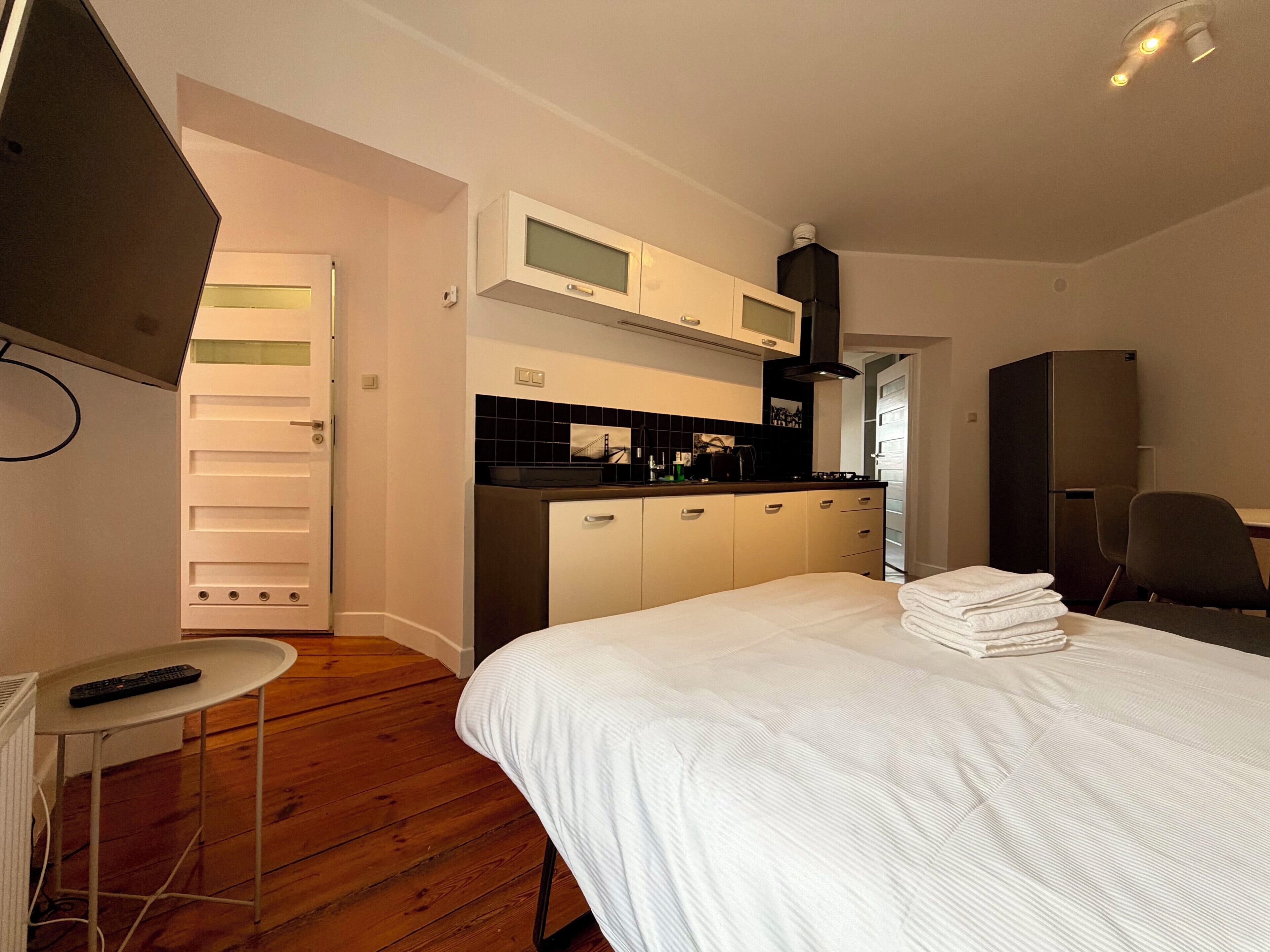 Un dormitorio con cama, una mesita, una silla, una zona de cocina con armarios y electrodomésticos, y una puerta que da a otra habitación.