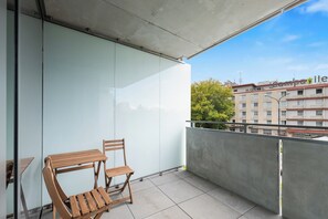 Un balcón con una barandilla de vidrio, una mesa y silla de madera, y vista a un edificio de hotel.