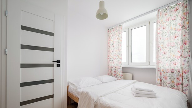 A bedroom with a bed, a window with floral curtains, a bedside lamp, and a door with horizontal stripes. - Puławska Urban Nest Apartment (Warszawa)